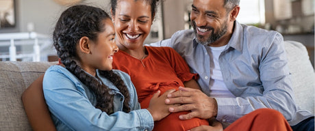 A pregnant woman sitting between a man and a child, their hands are on her stomach and they're all smiling.