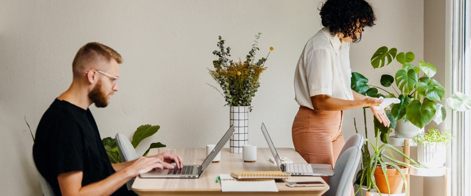 Sideview of a man working on a laptop on a table across from another open laptop while a woman tends to a plant in the corner.