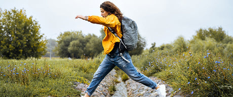 A woman with a backpack stepping over a stream in a field.