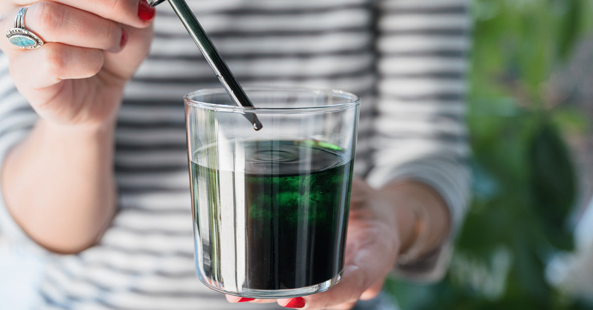 A woman adding liquid chlorophyll to a glass of water in her hand.