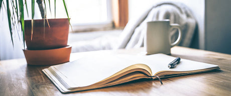 An open journal on a table with a pen and coffee cup.