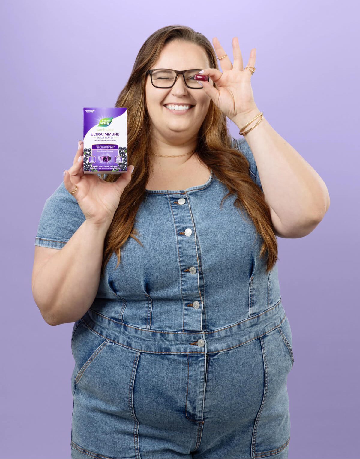 Woman in denim overalls holding Juicy Burst against a purple background