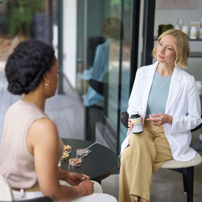 Woman in a white coat holding a supplement bottle talking to another woman in a modern office setting.