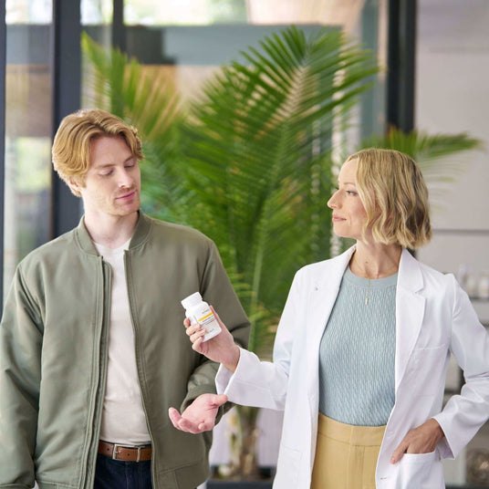 Woman in a white coat holding a bottle of pills to a man outdoors with plants in the background